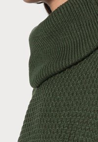 Grüner Strickpullover mit einem strukturierten Design und hohem, umgeschlagenem Kragen. Der Stoff hat ein geripptes, gewebtes Aussehen.