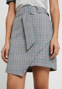 Grijze geruite rok met een riem met een ronde metalen gesp, diagonale wikkelontwerp en turquoise accenten. Gemaakt van lichte stof.