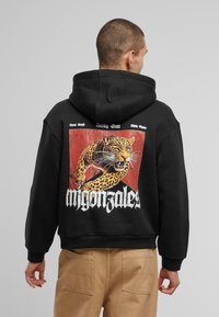 Schwarzer Kapuzenpullover aus weichem Stoff mit einem großen, bunten Leopardenmotiv auf dem Rücken und dem Text "migonza1es" in fetten weißen Buchstaben.