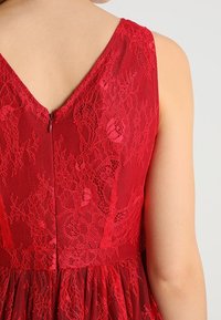 Vue arrière d'une personne portant une robe en dentelle rouge sans manches avec un décolleté en V et une fermeture éclair.