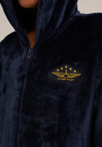 Navy blue, mīksts, ar rāvējslēdzēju apaļo kapuci, kas iezīmējas ar patīkamu tekstūru un dzeltenu "Nākotnes pilots" uzšuvumu ar spārnu emblēmu un zvaigznēm.