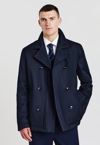 Cappotto navy doppiopetto realizzato in tessuto liscio, caratterizzato da grandi bottoni, colletto a intaglio e tasche laterali. Indossato sopra un completo.
