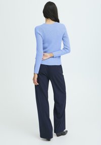Donna con lunghi capelli scuri che indossa un maglione a coste azzurro chiaro, pantaloni larghi blu scuro e scarpe con tacco nere, in piedi con la schiena rivolta verso la fotocamera.
