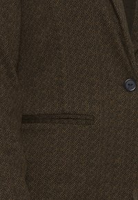 Blazer à motif chevrons marron et noir avec un bouton noir et une poche passepoilée horizontale sur le devant.