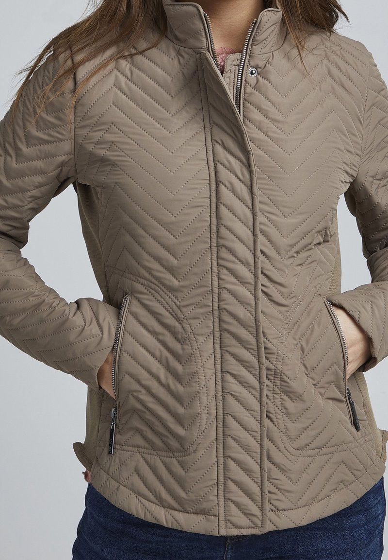 Jasnobrązowa pikowana kurtka w szewronowy wzór, z wysokim kołnierzem i bocznymi kieszeniami na zamek. Gładka faktura i dopasowany krój.