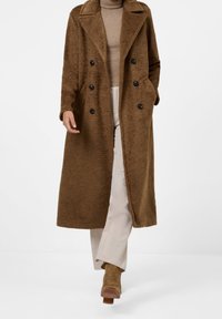 Brun, texturerad lång rock med dubbelknäppning och svarta knappar. Bärs över en beige polotröja och ljusa byxor, snyggt kombinerat med bruna stövlar.