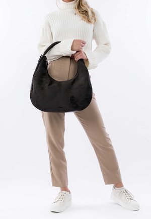 Sac hobo en daim noir avec une forme arrondie et une large bandoulière, porté par une personne vêtue d'un pull blanc à côtes et d'un pantalon beige.