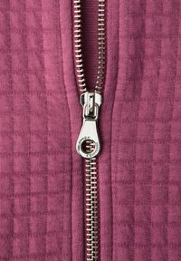 Detailní záběr stříbrného táhla zipu na částečně rozepnuté fialové texturované látce.