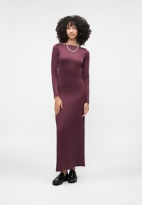 Duga haljina ljubičaste boje s dugim rukavima, izrađena od glatkog rastezljivog materijala s ravnom siluetom, u kombinaciji s crnim loafericama i necklace s biserima.