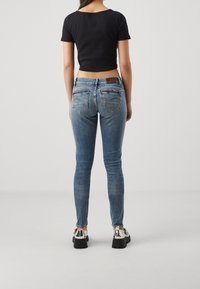 Jeans skinny azules con un lavado desgastado, dos bolsillos traseros y una etiqueta de cuero en la cintura. Combinados con una blusa negra ajustada y zapatos metálicos.