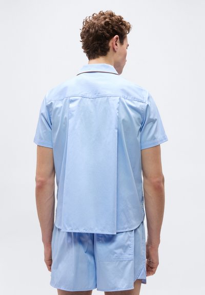 Άνδρας με σγουρά μαλλιά που φορά γαλάζιο κοντομάνικο πουκάμισο με γιακά και αντίστοιχο σορτς, εμφανιζόμενος από πίσω σε απλό φόντο.