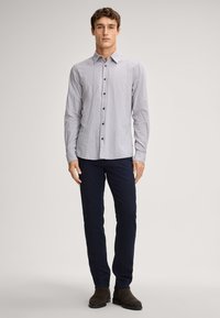 Camicia abbottonata in lino grigio chiaro con maniche lunghe, dotata di collo e bottoni marroni; indossata con pantaloni blu navy e scarpe marroni scure.