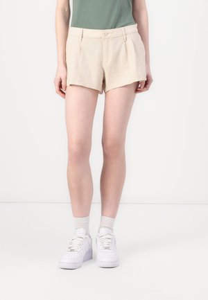 Persoon draagt beige, hooggetailleerde geplooide short met knoop en rits, gecombineerd met witte enkelsokken en witte sneakers.