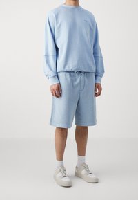 Man med ljusblå sweatshirt och matchande shorts, iförd vita sneakers och strumpor, på enfärgad bakgrund.