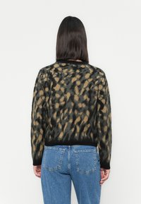 Mulher com cabelo liso e escuro veste um suéter de padrão leopardo preto e bege e jeans azuis, de costas para um fundo branco liso.