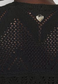 Nahaufnahme eines schwarzen Strickkleidungsstücks mit Diamant- und Lochmuster, verziert mit einem kleinen goldenen herzförmigen Twinset Milano Logo-Anhänger.