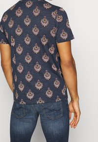 Mörkblå kortärmad skjorta med texturerad tyg, som har ett upprepande paisleymönster i livliga färger. Bärs med mörkblå jeans.