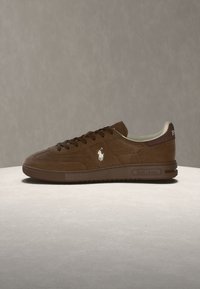 Καφέ χαμηλό sneaker με κορδόνια, λευκό λογότυπο αλόγου στη πλευρά και πιο σκούρα καφέ σόλα και ψηλοτάκουνο τμήμα, εμφανίζεται σε ανοιχτή επιφάνεια.