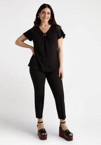 Bluza neagră cu decolteu în V și detaliu cu nod în față, asortată cu pantaloni negri ajustați și sandale cu talpă din lemn roșu-maro.