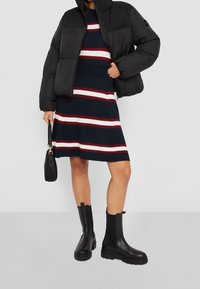 Chaqueta acolchada negra ajustada con un cuello alto. Vestido azul marino con rayas horizontales rojas y blancas. Botines gruesos negros y bolso.