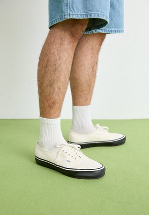 Sneakers bianchi in tela con suole in gomma nera, indossati con calzini bianchi a coste. Shorts di denim e pavimento verde sullo sfondo.