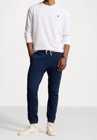 Camicia bianca a maniche lunghe con un piccolo logo blu navy, abbinata a pantaloni della tuta blu navy con tasche laterali e polsini elastici, e sneakers color avorio.