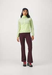 Giacca a zip verde chiaro con cappuccio, abbinata a pantaloni svasati di un rosso borgogna scuro e scarpe da trekking nere con dettagli rossi.