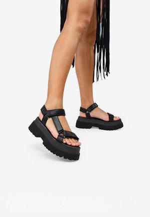 Sandales à plateforme noires avec semelles épaisses, larges brides et finition mate. Le design comprend un bout ouvert et une bride de cheville ajustable.