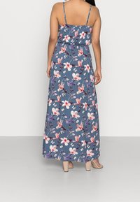 Kvinna som bär en blå blommig maxiklänning med tunna band, sedd bakifrån, stående på ett grått golv med en vit bakgrund.