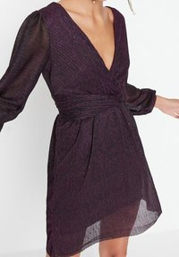 Fioletowa sukienka z długimi, bufiastymi rękawami i głębokim dekoltem w kształcie V. Posiada ściągniętą talię i zwiewny dół, wykonana z teksturowanej, tkanej tkaniny.