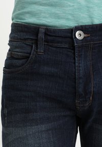 Jeans de mezclilla azul oscuro con cintura ajustada, bolsillos frontales y cierre de botón plateado. La tela tiene una textura suave y un ligero desgaste.