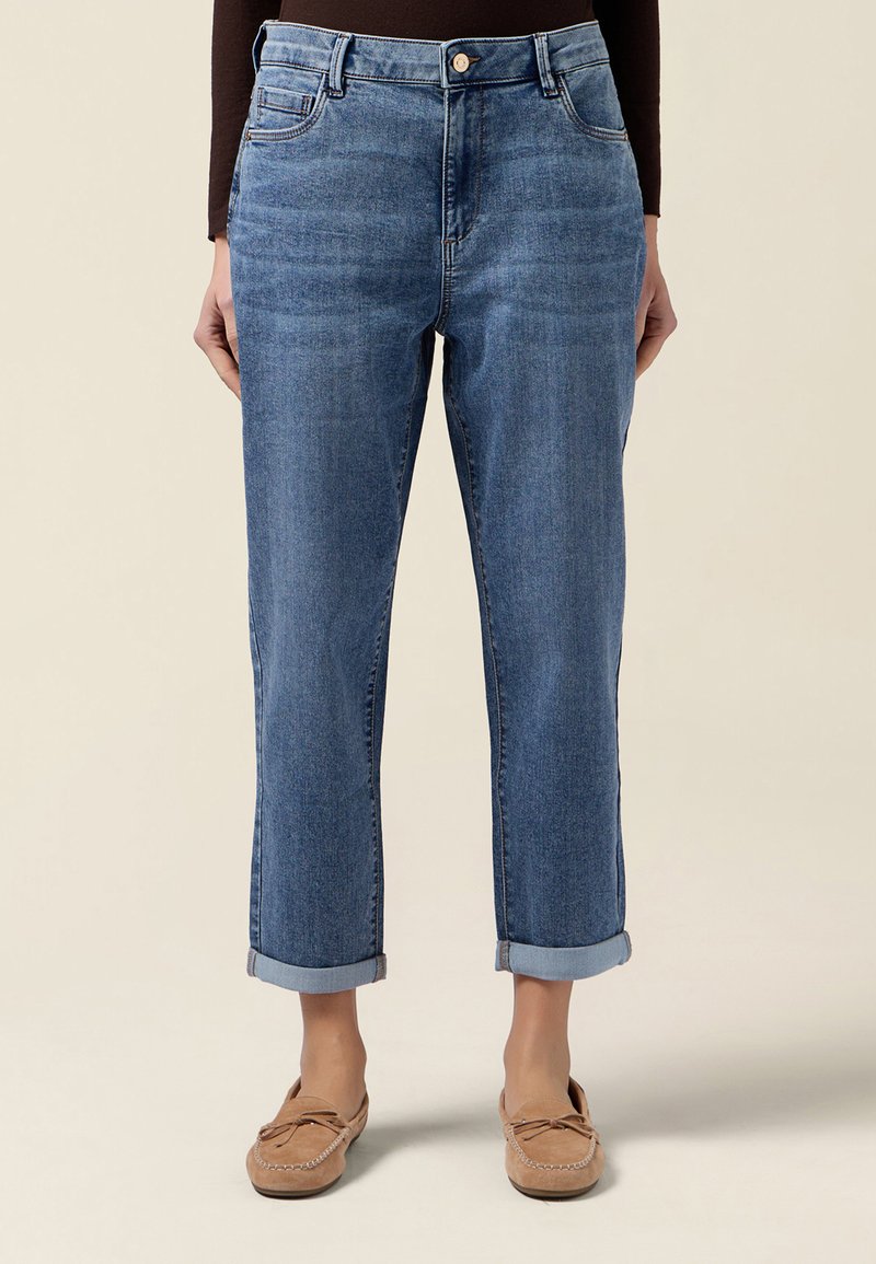 Niebieskie dżinsy z prostą nogawką i mankietami, noszone z brązowymi mokasynami i ciemnym topem z długim rękawem, pokazane od talii do stóp na jasnym tle.