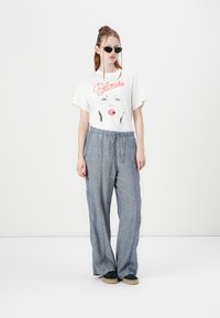 Hvid t-shirt med sort og rød grafisk tryk, parret med løstsiddende grå bukser og sorte slip-ons med en beige sål.