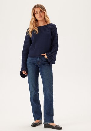 Maglione blu navy lavorato a maglia con texture a coste sulle maniche, abbinato a jeans blu a gamba dritta e ballerine nere.