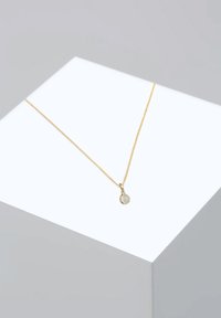 Gouden halsketting met een fijne ketting en een cirkelvormige hanger met een doorschijnende edelsteen, tentoongesteld op een geometrische sokkel.