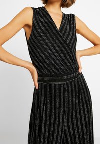 Robe noire sans manches avec des rayures verticales argentées, décolleté croisé, et taille cintrée portée par une personne les mains sur les hanches.