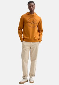 Felpa arancione con una stampa grafica sul petto, abbinata a pantaloni beige e scarpe da ginnastica bianche con dettagli marrone chiaro.