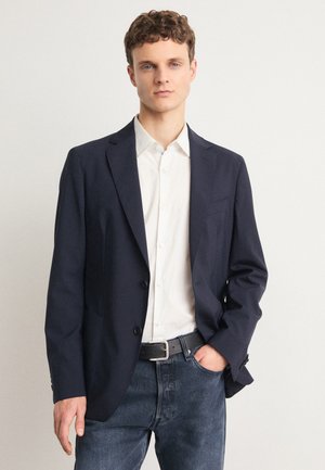 Navy blazer with a two-button front, tailored fit, and notch lapels. Worn over a white shirt, paired with blue denim jeans.