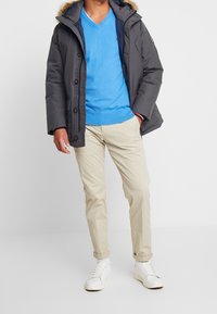 Graue Pufferjacke mit einer mit Fell gefütterten Kapuze, blauer Sweater, beiger Chinohose und weißen Sneakers. Verfügt über Knöpfe und kontrastierende Akzente.