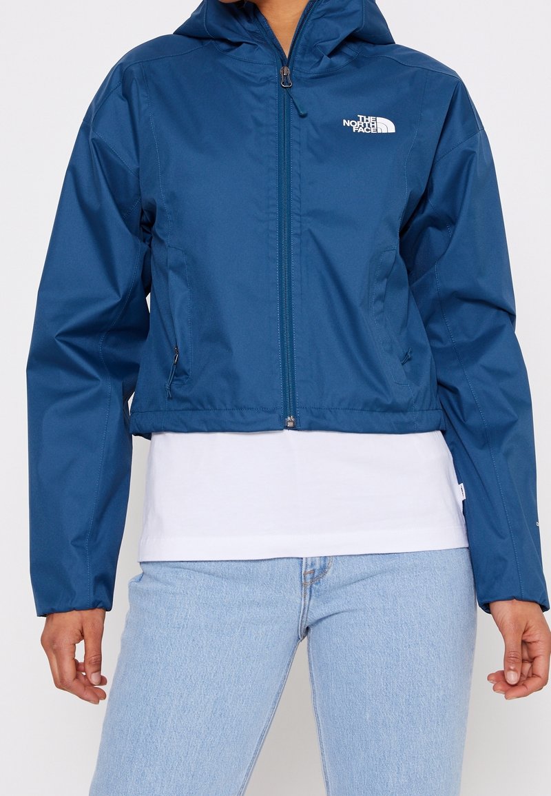 Femme portant une veste courte zippée North Face bleue avec capuche, chemise blanche en dessous, et jean bleu clair sur un fond uni.