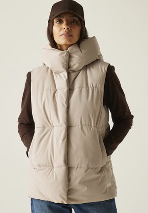 Kobieta w beżowej pikowanej kamizelce z wysokim kołnierzem, ciemnej koszulce z długim rękawem, niebieskich dżinsach i ciemnobrązowej czapce, stojąca z rękami w kieszeniach.