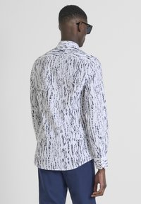Camicia bianca con motivi astratti neri, maniche lunghe, colletto a punta e orlo curvo, abbinata a pantaloni blu navy.