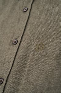 Olive green button-up shirt made of textured fabric with a herringbone pattern. Features dark buttons and an embroidered logo on the pocket.