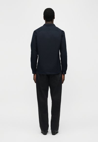 Άνδρας που στέκεται με την πλάτη προς τα εμπρός, φοράει μακρυμάνικο μπλε σκούρο πουκάμισο και άνετα μαύρα τζιν, μπροστά σε απλό φωτεινό φόντο.