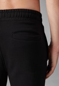 Schwarze Sweatpants haben einen breiten, elastischen Bund und eine kleine, schlanke Gesäßtasche. Der Stoff wirkt glatt und flexibel.