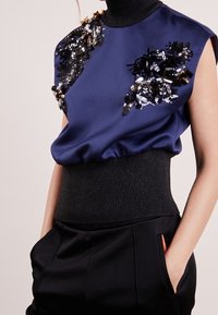 Marinblus i satin med dekorerade axlar som har paljetter och texturer, ribbad svart midja, matchad med svarta högmidjade byxor.