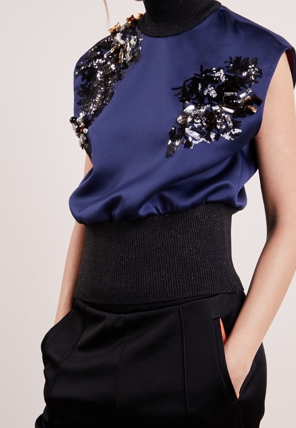 Marinblus i satin med dekorerade axlar som har paljetter och texturer, ribbad svart midja, matchad med svarta högmidjade byxor.