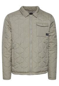 Veste matelassée beige clair avec col, fermeture éclair frontale complète et poche poitrine gauche ; présente une texture lisse et un motif discret gonflé.