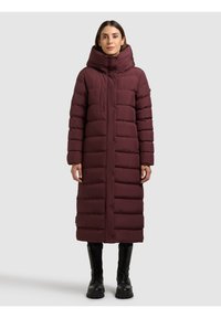 Hosszú, bordó pufi kabát magas gallérral, vízszintes steppelt mintával és elől patentgombokkal, fekete fűzős csizmával párosítva.