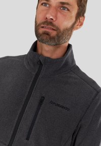 Grijze fleece jas met hoge kraag, voorzien van een zwarte ritsborstzak en geborduurd logo. Zachte textuur met minimale ontwerpelementen.